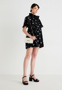 Black dress with white bow pattern, ruffled collar and sleeves. Pair of black high-heeled sandals. White handbag with chain strap.
