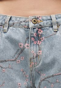 Ανοιχτό μπλε τζιν παντελόνι με ορειχάλκινο κουμπί και σχέδιο ροζ ανθισμένου κλαδιού κοντά στη μέση και στην περιοχή του φερμουάρ.