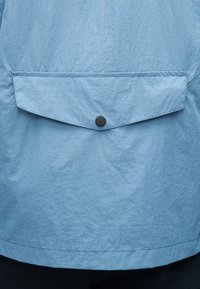 Tecido de casaco azul claro com superfície texturada, apresentando um bolso de aba com um botão de pressão preto na parte traseira.