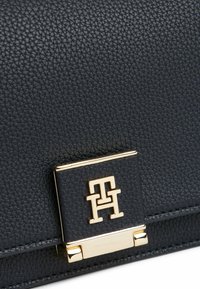 Sac en cuir noir texturé avec un fermoir doré gravé du logo. Design rectangulaire compact avec des détails de couture visibles.