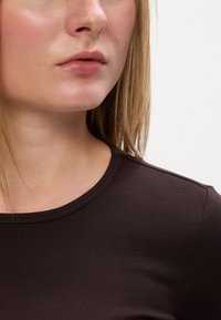 Hnedý priliehavý top s okrúhlym výstrihom. Hladká, jemná tkanina. Detailný záber na krk a dolnú časť tváre, so stredne dlhými blond vlasmi.