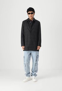 Abrigo de cuadros negros con un corte relajado, cierre de botones y bolsillos laterales, combinado con pantalones cargo de mezclilla azul claro y zapatillas blancas.