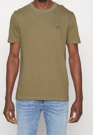 Man draagt een olijfgroen T-shirt met korte mouwen en ronde hals met een klein vierkant logo op de borst, gecombineerd met lichtblauwe jeans.