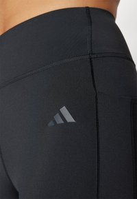 Leggings sportivi neri realizzati in tessuto elasticizzato. Presentano cuciture piatte, una vita alta e un logo Adidas riflettente sul lato.