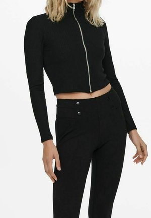 Femme portant une veste courte noire ajustée à manches longues avec fermeture éclair devant et un pantalon noir taille haute sur fond blanc.