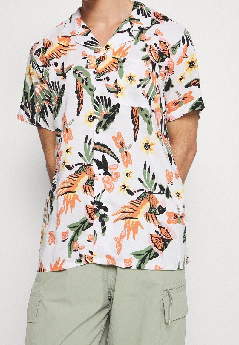 Chemise blanche à manches courtes boutonnée avec des imprimés colorés de poissons tropicaux, papillons et fleurs, portée avec un pantalon cargo vert clair.