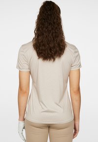 Top beige a maniche corte con un sottile motivo geometrico, caratterizzato da una vestibilità rilassata e una superficie liscia, indossato con pantaloni chiari e guanti.