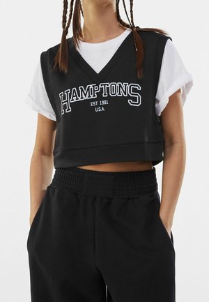 Frau trägt ein schwarzes ärmelloses Crop-Top mit dem Schriftzug "HAMPTONS EST 1991 U.S.A." über einem weißen, hochgekrempelten kurzärmligen Shirt und schwarzen Hosen.