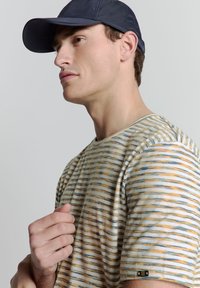 T-shirt rayé en beige, orange et bleu ; col rond ; texture douce ; manches courtes ; petit accent noir sur la manche.
