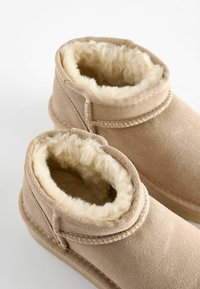Beige Knöchelschuh aus weichem Wildleder mit einem plüschigen, cremefarbenen Lammfellfutter. Merkmale sind verstärkte Nähte und eine flexible Gummisohle.