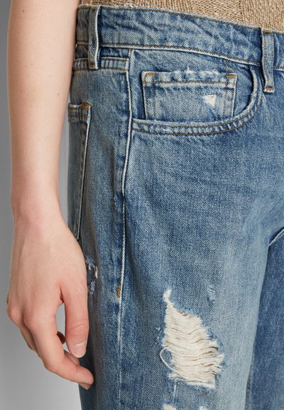 Jeans de denim desgastado em azul claro com remendos desfiados, costura visível e design clássico de cinco bolsos; mão descansando na perna da calça.