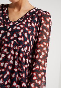 Laivastonsininen pusero, jossa on valkoisia ja punaisia kukkakuvioita, läpinäkyvät hihat, V-pääntie ja pitsireunus. Mukana on kerätty vyötärö ja kevyt kangas.