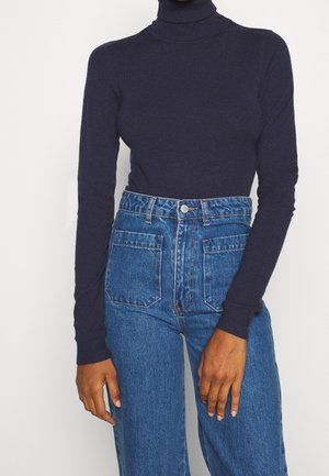 Person iført en tætsiddende navy rullekravesweater stoppet ned i højtaljede blå jeans med forlommer, stående mod en ensfarvet baggrund.