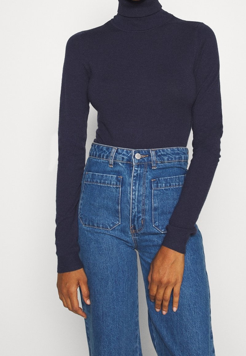 Personne portant un pull à col roulé bleu marine ajusté, rentré dans un jean bleu taille haute avec des poches plaquées à l'avant, debout devant un fond uni.