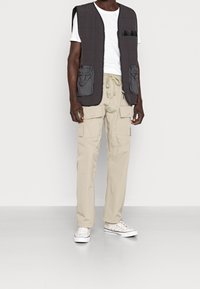 Prošívaná šedá bezrukávová vesta se dvěma předními kapsami, zkombinována s béžovými cargo kalhotami s několika kapsami a stahovacími šňůrkami v pase. Bílé tenisky.