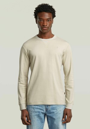 Jonge man met kort krullend haar, gekleed in een lichtbeige shirt met lange mouwen en lichtblauwe spijkerbroek, staand tegen een effen achtergrond.