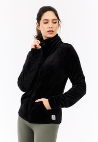 Schwarze Fleecejacke mit hohem Kragen, durchgehendem Reißverschluss und Seitentaschen. Glatte Textur mit einem Markenschild am Saum.