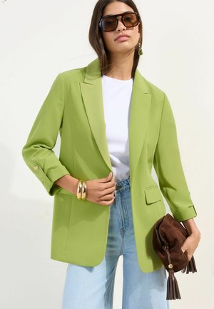 Femme portant un blazer vert citron surdimensionné, une chemise blanche, un jean bleu clair, de grandes lunettes de soleil, des bracelets en or, tenant une pochette en cuir marron avec des pampilles.