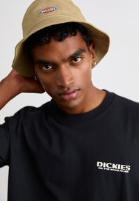 Maglietta nera con testo bianco "DICKIES ON THE ROAD AGAIN" e cappello a secchiello beige con logo ricamato. Modello maschile giovane con capelli ricci.