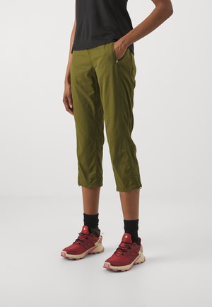 Person steht und trägt olivgrüne kurze Hosen mit Reißverschlusstaschen, schwarze Socken, rote Trailrunning-Schuhe und ein schwarzes Oberteil, Hände in den Taschen.