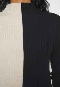 Kvinna som bär en ribbad långärmad polotröja med horisontell färgblockering i halva beige och halva svart.