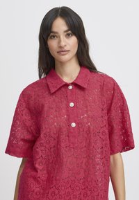 Camicia rossa in pizzo con maniche corte, dotata di colletto e tre bottoni perla. Un intricata fantasia floreale aggiunge texture e dettaglio.