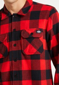 Camisa de franela de cuadros rojos y negros con cierre de botones en la parte frontal, dos bolsillos en el pecho y una etiqueta de tela en el bolsillo izquierdo. Apariencia texturizada.