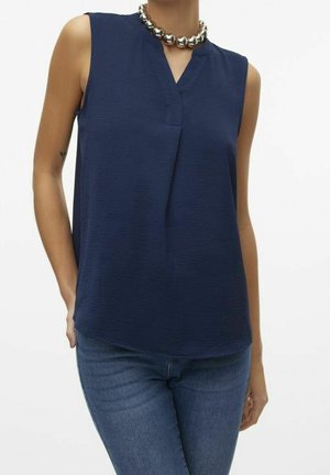 Kobieta ubrana w bezrękawnik o teksturowanej, granatowej bluzce, srebrny masywny naszyjnik i niebieskie dżinsy, stojąca z jedną ręką w kieszeni.