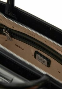 Interno di borsa sintetica nera con fodera beige. Presenta una tasca con cerniera, una targhetta con il logo e un motivo ripetuto "GUESS". Ferramenta color argento.