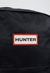 Gros plan d’une surface en tissu noir avec une étiquette rectangulaire blanche bordée de rouge affichant le mot "HUNTER" en lettres noires.
