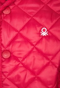 Prošívaná červená bunda s kulatými knoflíky a bílým vyšívaným logem chobotnice na hrudi. Hladká syntetická tkanina s lesklým povrchem.