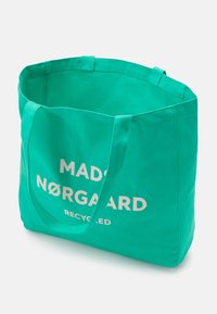 Újrahasznosított szövet bevásárlótáska élénk türkiz színben. Két strapabíró fogantyúval rendelkezik, és fehér nyomtatott szöveggel "MADØ NØRGAAARD RECYCLED".
