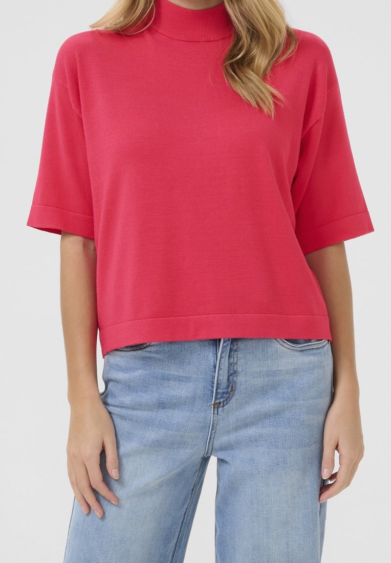 Femme portant un pull rose vif à manches courtes avec un col montant et un jean bleu clair, vue des épaules aux hanches, debout immobile.