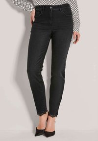 Vrouw in zwarte slim-fit jeans met kantafwerking bij de enkels, gecombineerd met zwarte pointe-heeled schoenen en een geblokte blouse met lange mouwen.