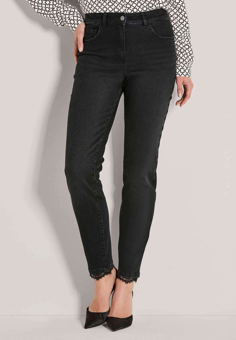 Vrouw in zwarte slim-fit jeans met kantafwerking bij de enkels, gecombineerd met zwarte pointe-heeled schoenen en een geblokte blouse met lange mouwen.