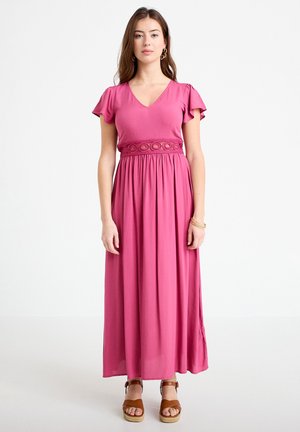 Femme debout portant une robe maxi rose à col en V, manches évasées, détail décoratif à la taille, et sandales compensées marron.