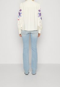 Vit långärmad blus med färgglad blommig brodyr på ärmarna, kombinerad med ljusblå utsvängda jeans. Slätt tyg med en lätt lyster.