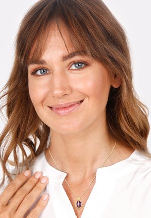 Goldene Halskette mit einem lila ovalen Anhänger an einer dünnen Kette. Der Anhänger hat dekorative Akzente. Modell trägt eine weiße Bluse, sanft beleuchteter Hintergrund.