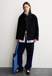 Schwarzer Wollmantel mit großen Knöpfen, über ein gestreiftes Hemd getragen. Dunkelblaue Cordhosen und eine blaue Tragetasche vervollständigen das Outfit.