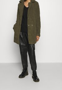 Persona vestida con un abrigo texturizado verde oliva, suéter negro de cuello en V, pantalones de cuero negros y botas negras con cordones, de pie contra un fondo liso.