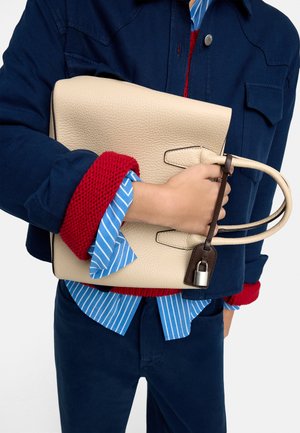 Osoba ubrana w warstwowe niebiesko-czerwone ubranie trzyma beżową torebkę o teksturowanej powierzchni z podwójnymi uchwytami i srebrną kłódką.