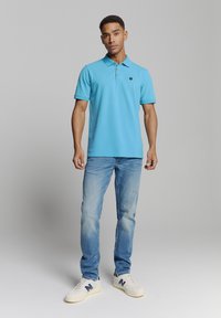 Lichtblauwe polo met kraag en drieknops sluiting, gecombineerd met lichtblauwe jeans en witte sneakers met een marineblauw logo.
