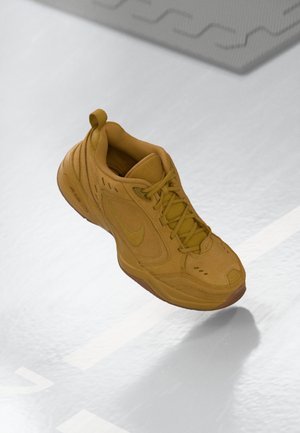 Une seule chaussure Nike jaune moutarde à lacets flottant au-dessus d’un sol gris clair avec des marques blanches et un bord de tapis à motif visible.
