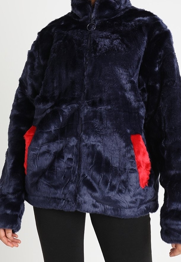 Femme portant une veste zippée en peluche bleu marine avec des poches en peluche rouge vif, associée à un pantalon noir.