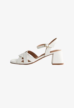 Witte leren sandaal met open teen, gekruiste banden, enkelgesp en blokhak op een witte achtergrond.