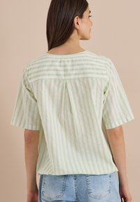 Femme aux longs cheveux bruns porte une chemise blanche à manches courtes avec des rayures verticales vert clair et un jean bleu clair, vue de dos.