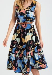 Robe florale avec un fond noir ornée de fleurs colorées, à une épaule, jupe à volants et ceinture tressée marron.