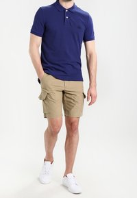 Polo blu navy con maniche corte e due bottoni; abbinato a pantaloni cargo khaki con tasche laterali; sneakers bianche.