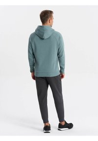 Sudadera verde con un bolsillo canguro frontal, puños y dobladillo de canalé. Combinada con joggers grises ajustados y zapatillas negras.
