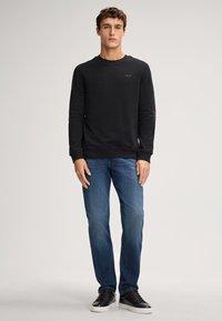 Felpa nera con collo girocollo, tessuto testurizzato, abbinata a jeans in denim blu. Il modello indossa sneakers nere, con le mani sui fianchi.